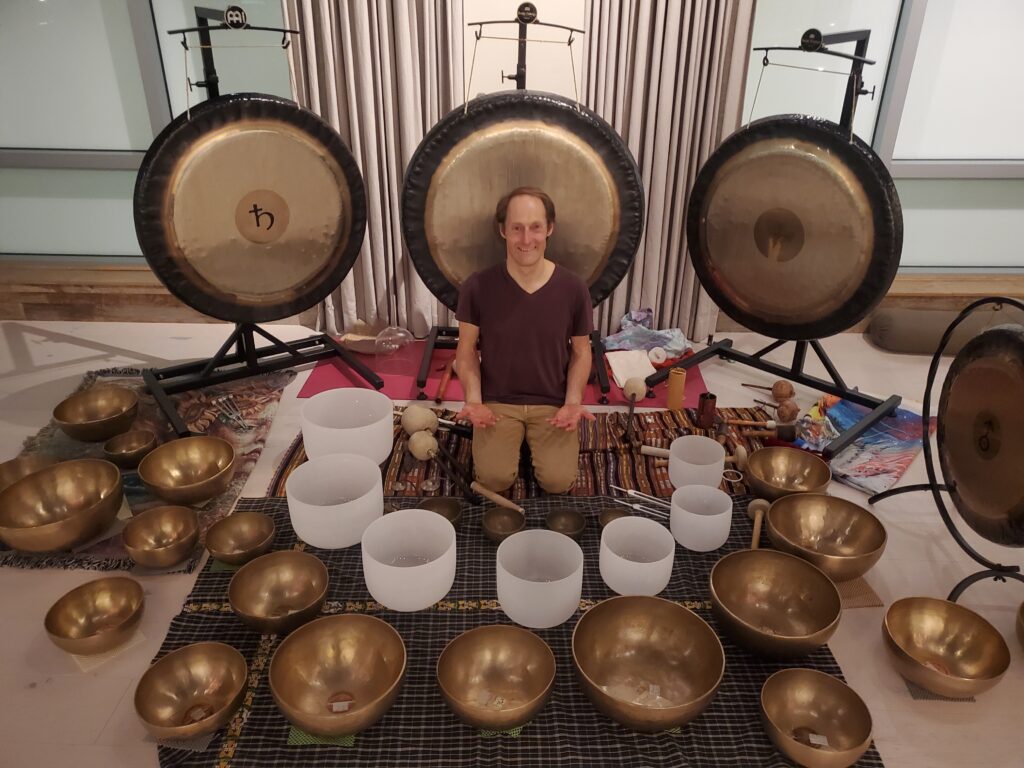 Kirk seated with smile. 4 gongs behind him, 7 crystal bowls in front, and dozens of bronze bowls